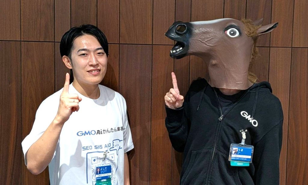 Tシャツ姿の男性と馬のかぶりものをした人物が、指を立てて並んで立っている写真